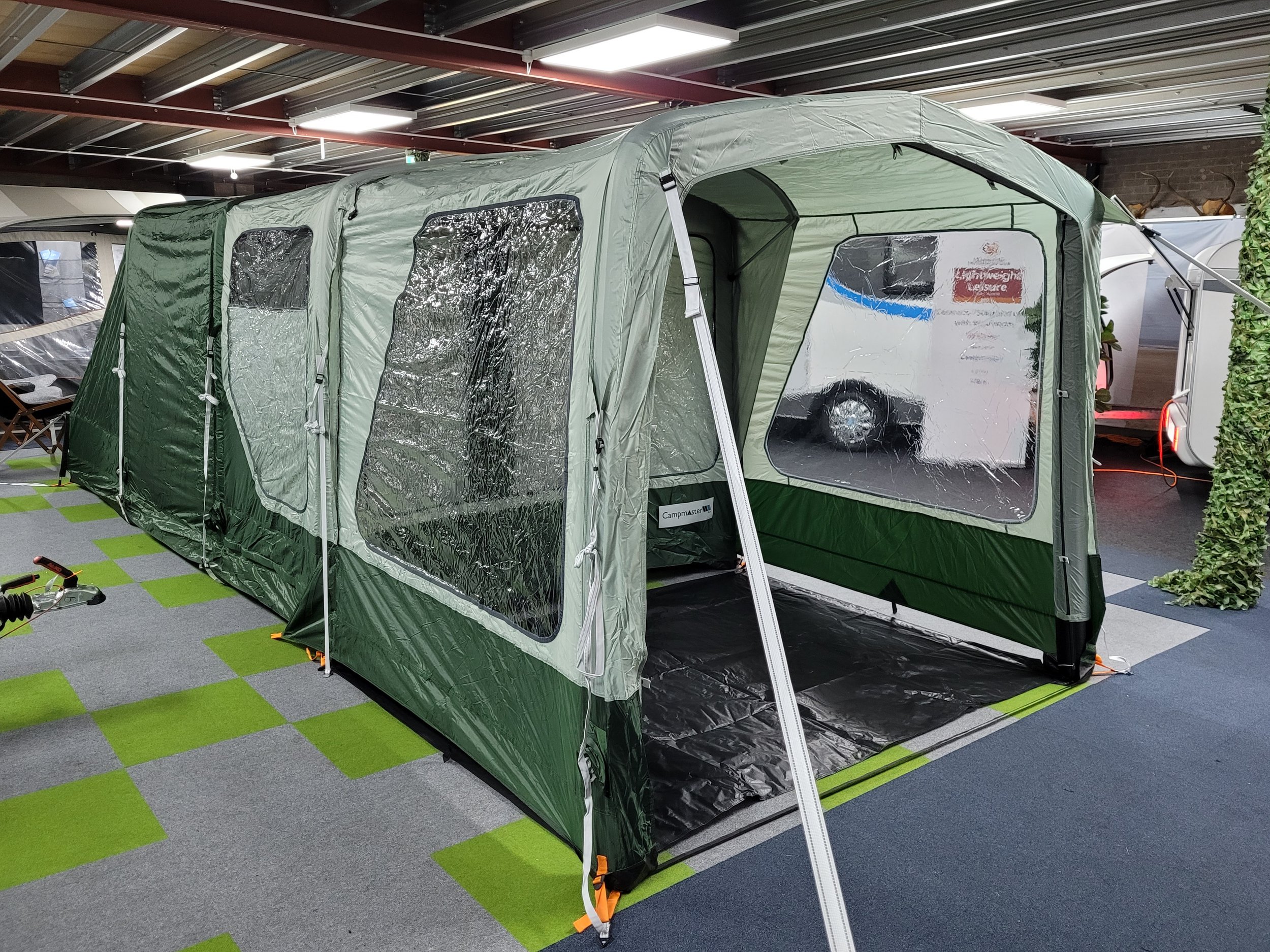 Campmaster Air A large green camping tent with transparent windows on display indoors, set up on a checkered green and gray carpet with additional camping gear around.
