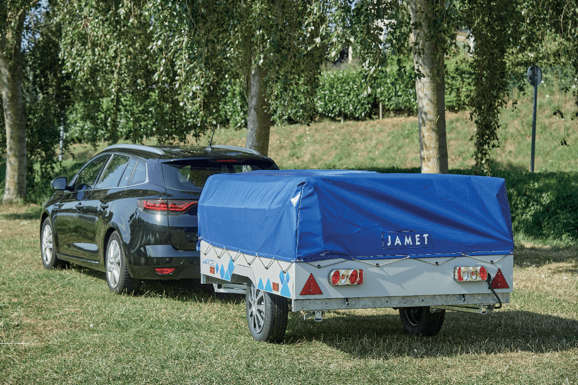 Black car with a blue covered Jamet trailer attached, parked on grass near trees and a hill.