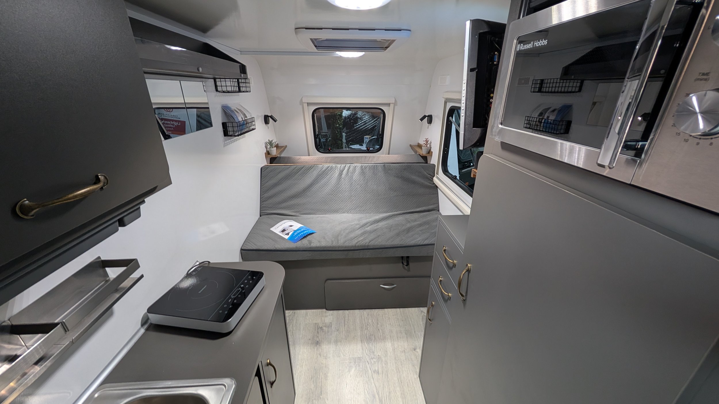 Interior of a compact modern camper van with a bed, kitchen appliances, and storage cabinets, viewed from the kitchen area looking toward the back window.
