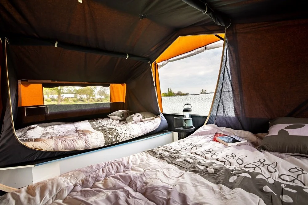 Inside a large Camplair with two beds; one has a view outside through a window, and there's a nightstand with a lantern and mug between the beds.