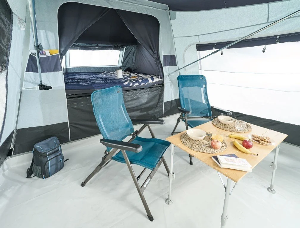 Interior of a spacious Jamet Compact camping tent with a bed, two blue chairs, a wooden table set with bowls, bananas, apples, a book, and a small backpack.
