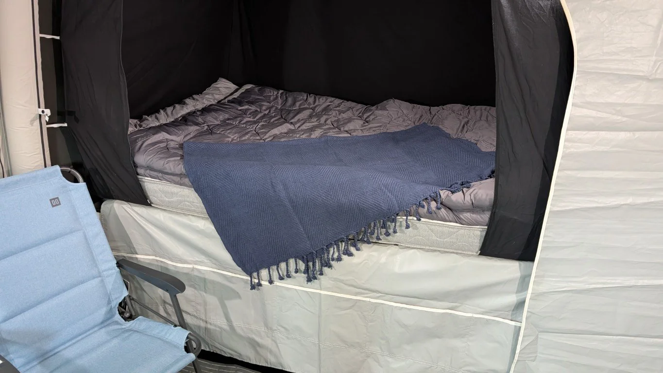 A bed inside a pop-up camper with grey bedding and a blue throw blanket. The camper has black curtains around the bed.