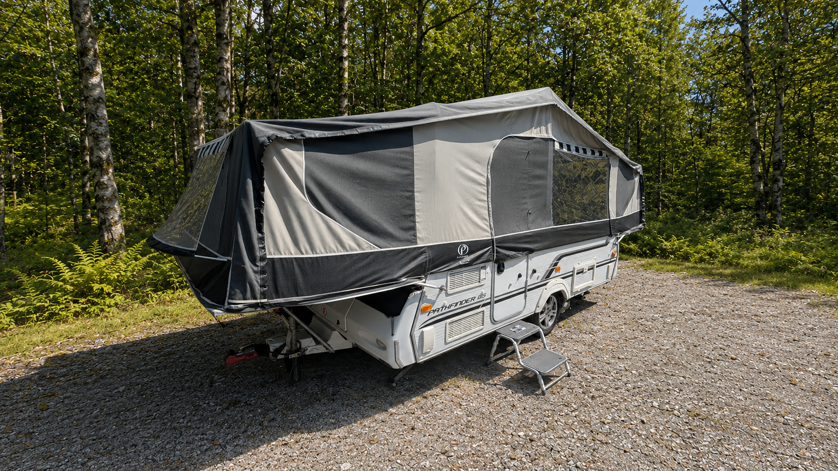 A white travel trailer with a covered awning set up on a gravel parking area in a wooded area with green trees and ferns in the background.