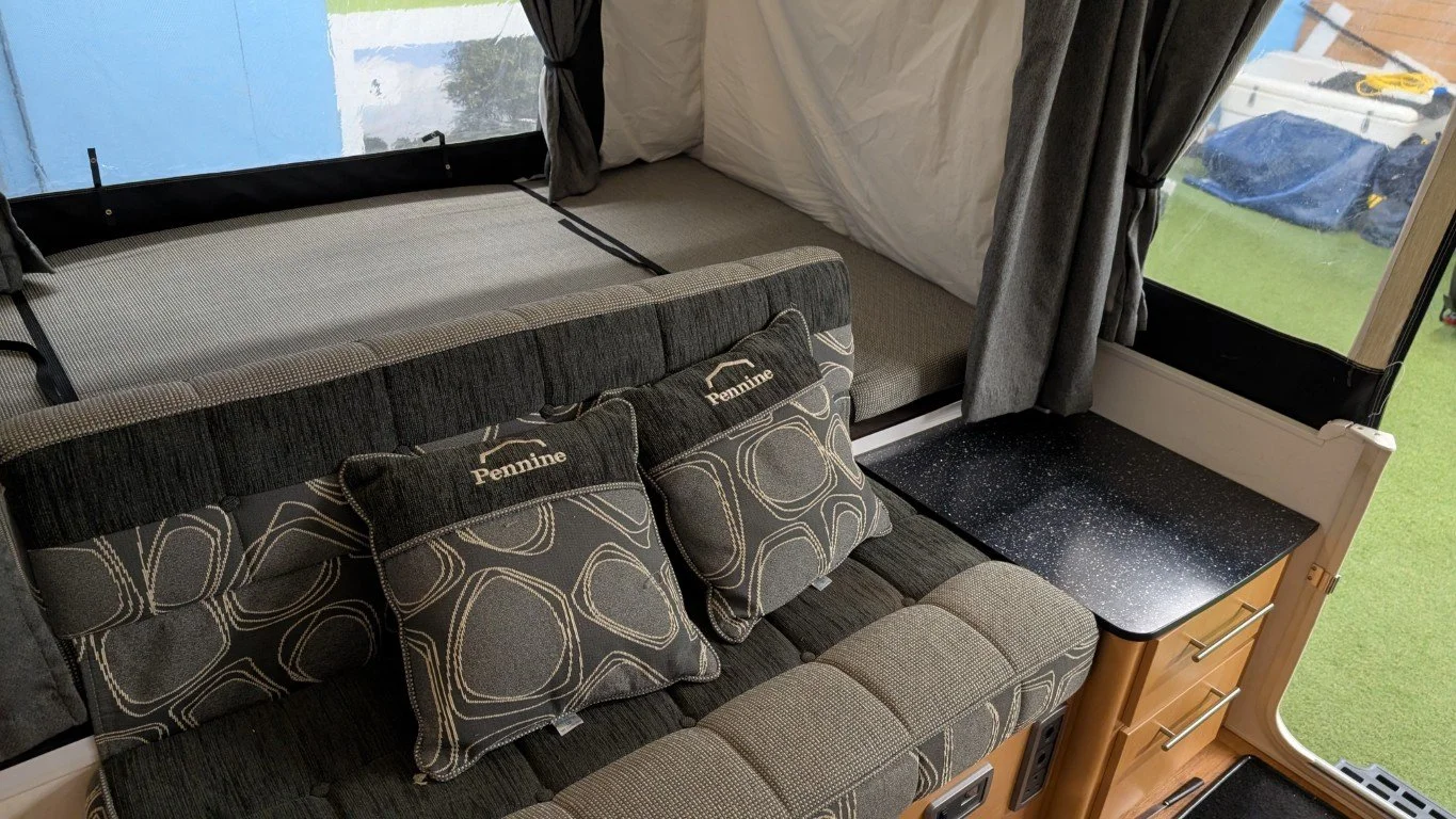 Inside a camper, with a sofa and a Murphy bed with a fold-up mattress, window with curtains, and a small countertop next to the door.
