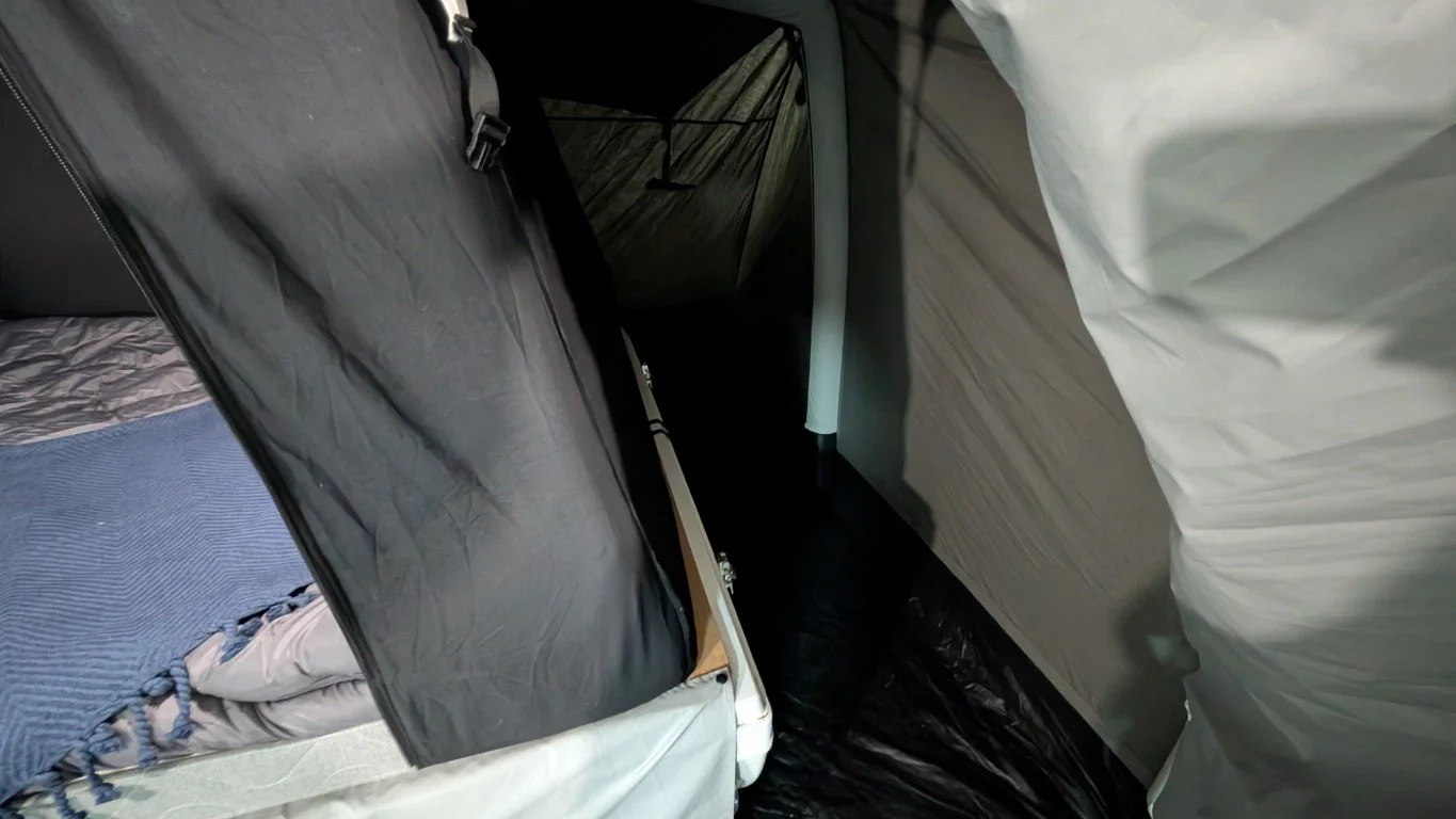 Inside a tent showing a narrow corridor with a bed partially visible on the left and hanging fabric or tent wall panels on the sides.