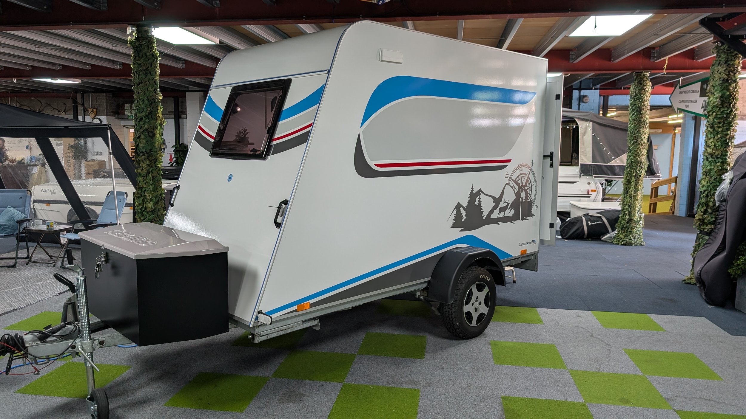 A white travel trailer with blue, red, and gray graphics displayed indoors at a showroom. The trailer has a black front storage compartment, a side window, and is surrounded by other campers and equipment on a checkered green and gray floor with deco