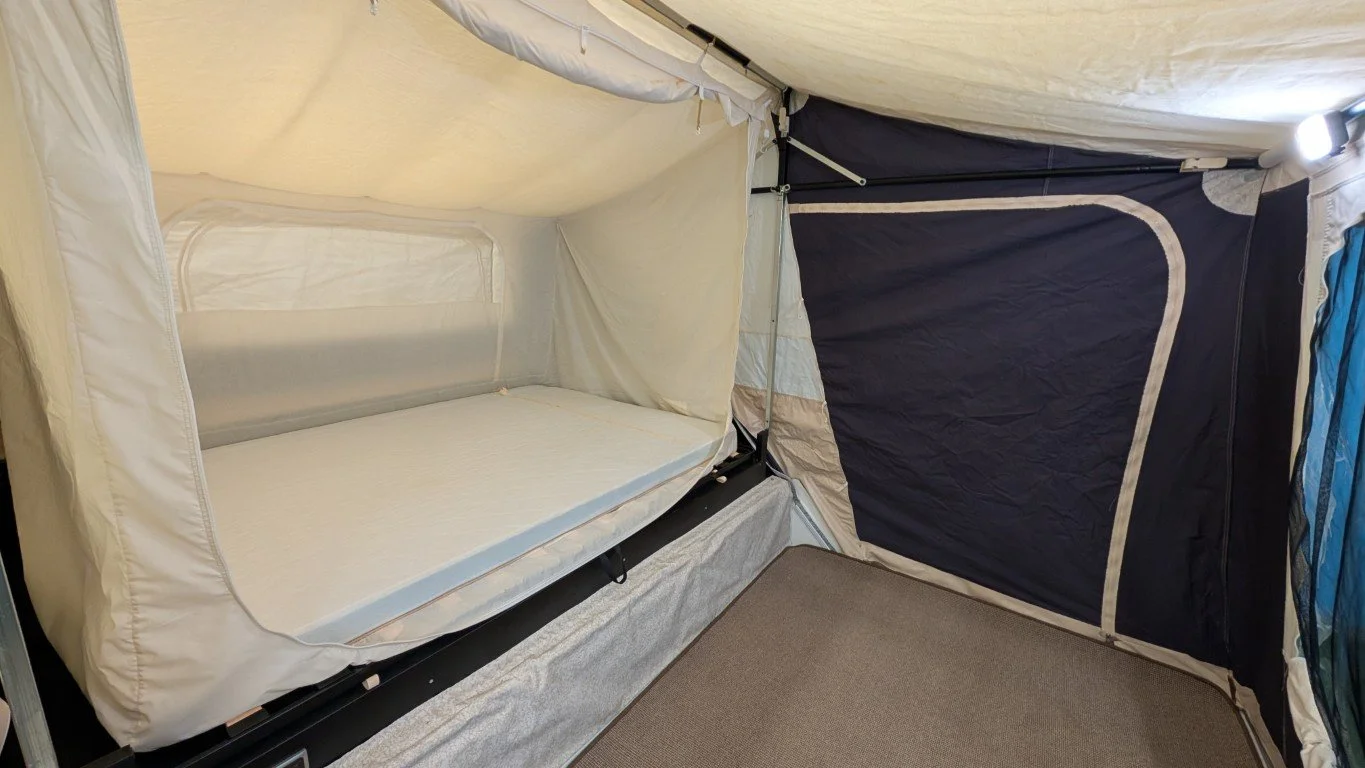 The image shows the interior of a tent with a built-in sleeping area featuring a mattress and a tent window, with a side wall made from blue material.