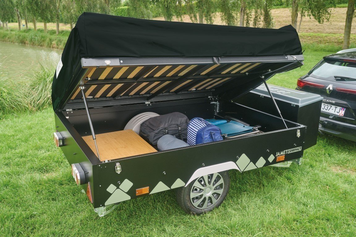 A small black Jamet camping trailer with an open lid, revealing storage compartments and camping bags inside, parked on grass near a pond and trees.