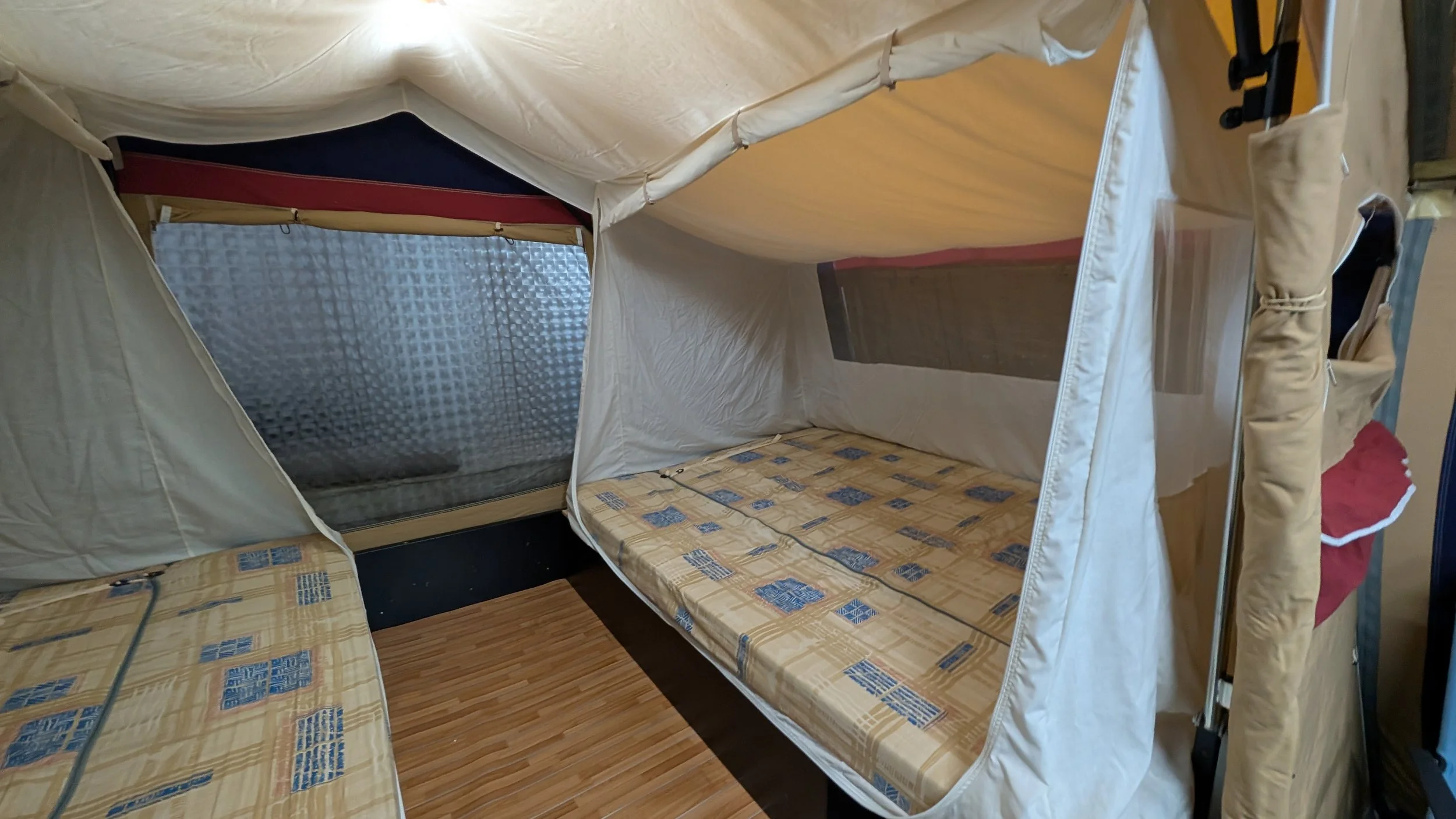 Inside a pop-up camping tent with two sleeping areas and a window, set up indoors on a wooden floor.
