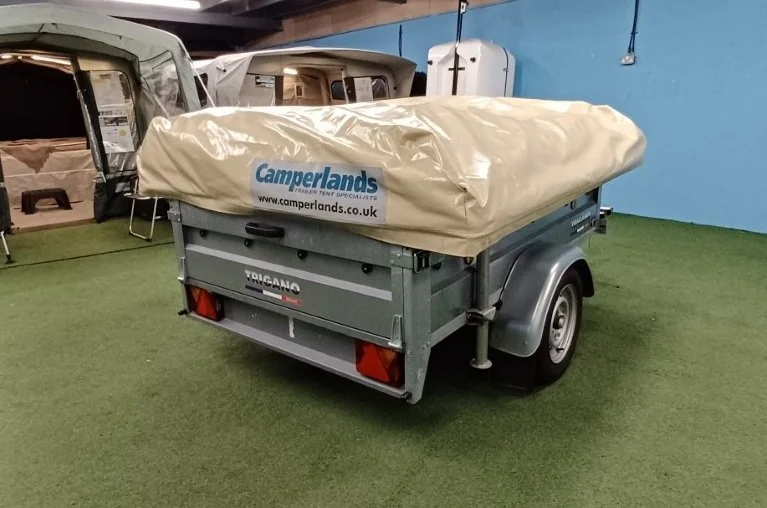 A small gray portable trailer with a tan cover labeled "Camperlands" is parked indoors on green artificial grass, with other tents or trailers visible in the background.