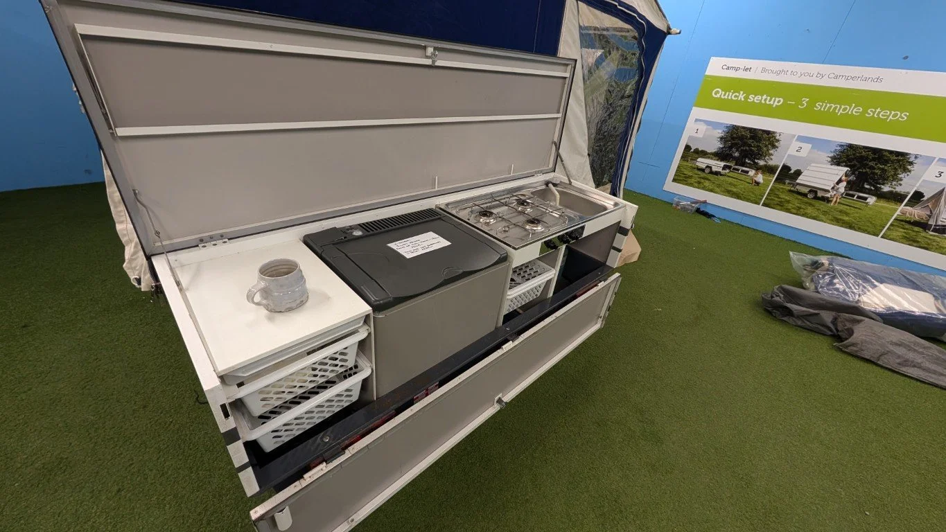Portable camping kitchen setup with a stovetop, sink, and storage, on green artificial grass with a blue wall and an informational poster in the background.