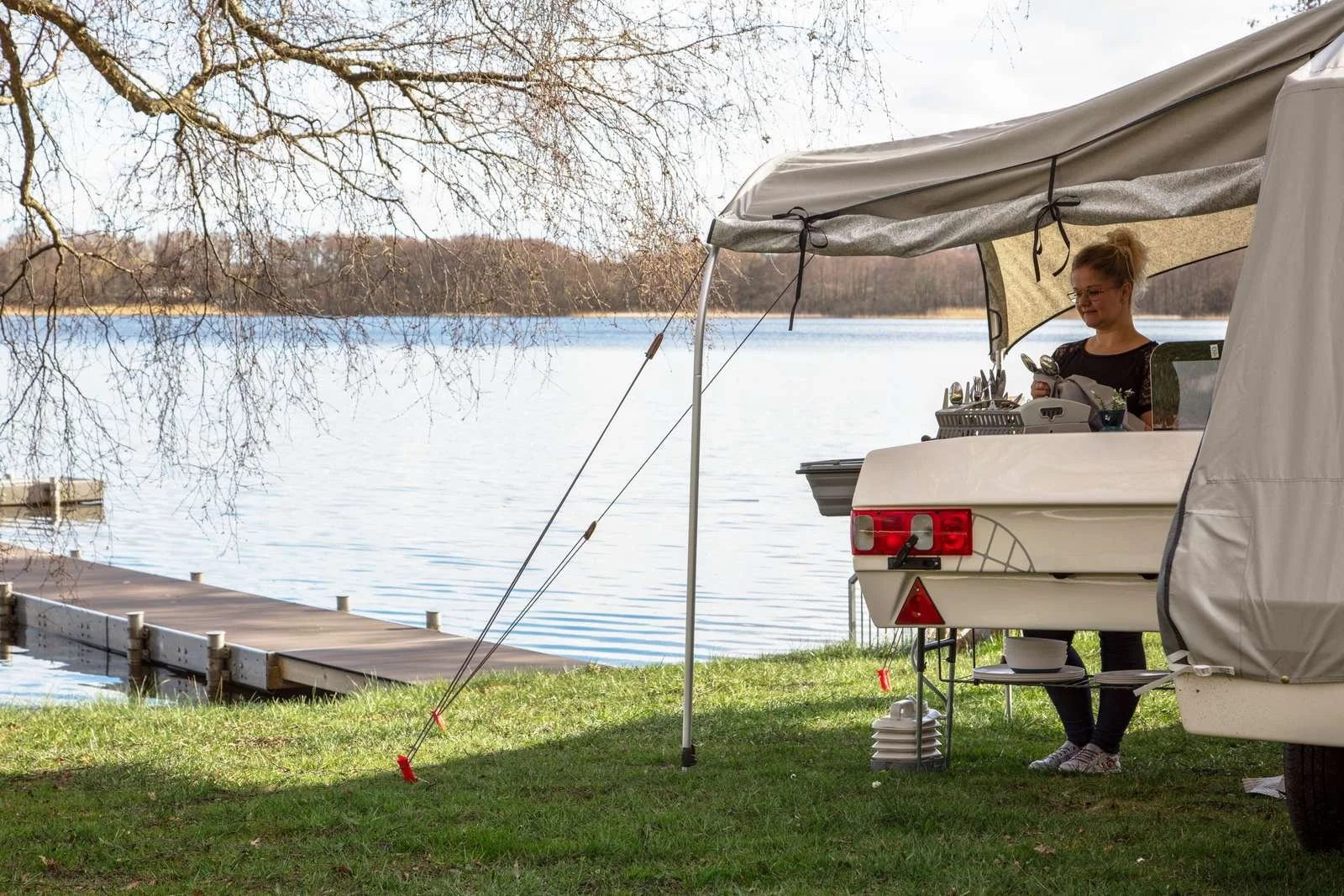 A woman standing inside a Camp-let by a lakeside, using a smartphone, with a wooden dock extending into the lake and leafless trees in the background.