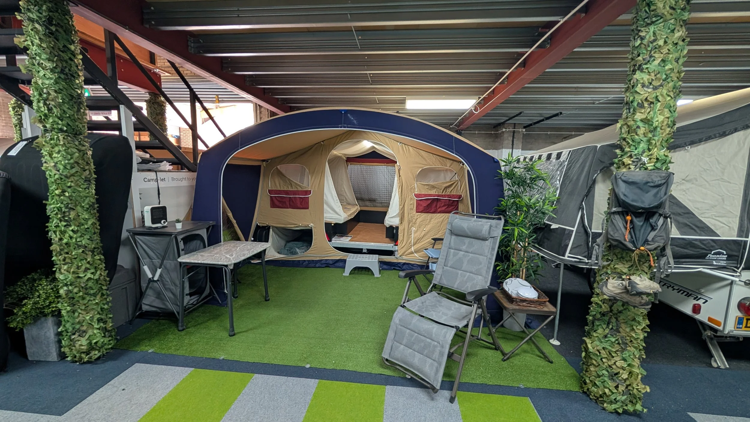 Indoor showroom with camping tents, chairs, and outdoor gear on artificial grass and colorful carpet tiles.