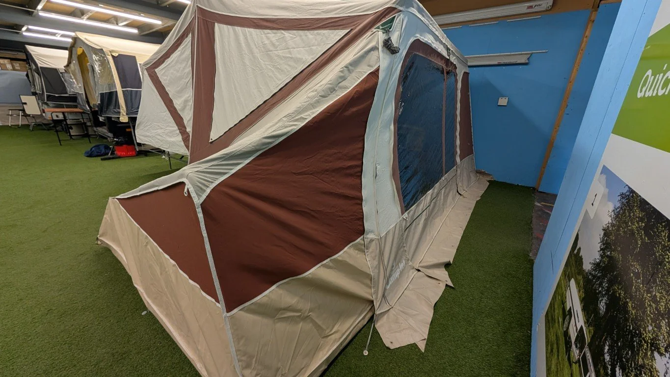 A military-style tent with brown and beige fabric, large windows with mesh screens, is set up indoors on green artificial turf, surrounded by other tents and displays.
