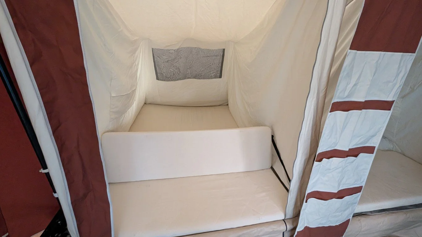 Empty hotel bunk bed with cream-colored mattress and safety rail, inside a fabric privacy tent with red and white stripes.