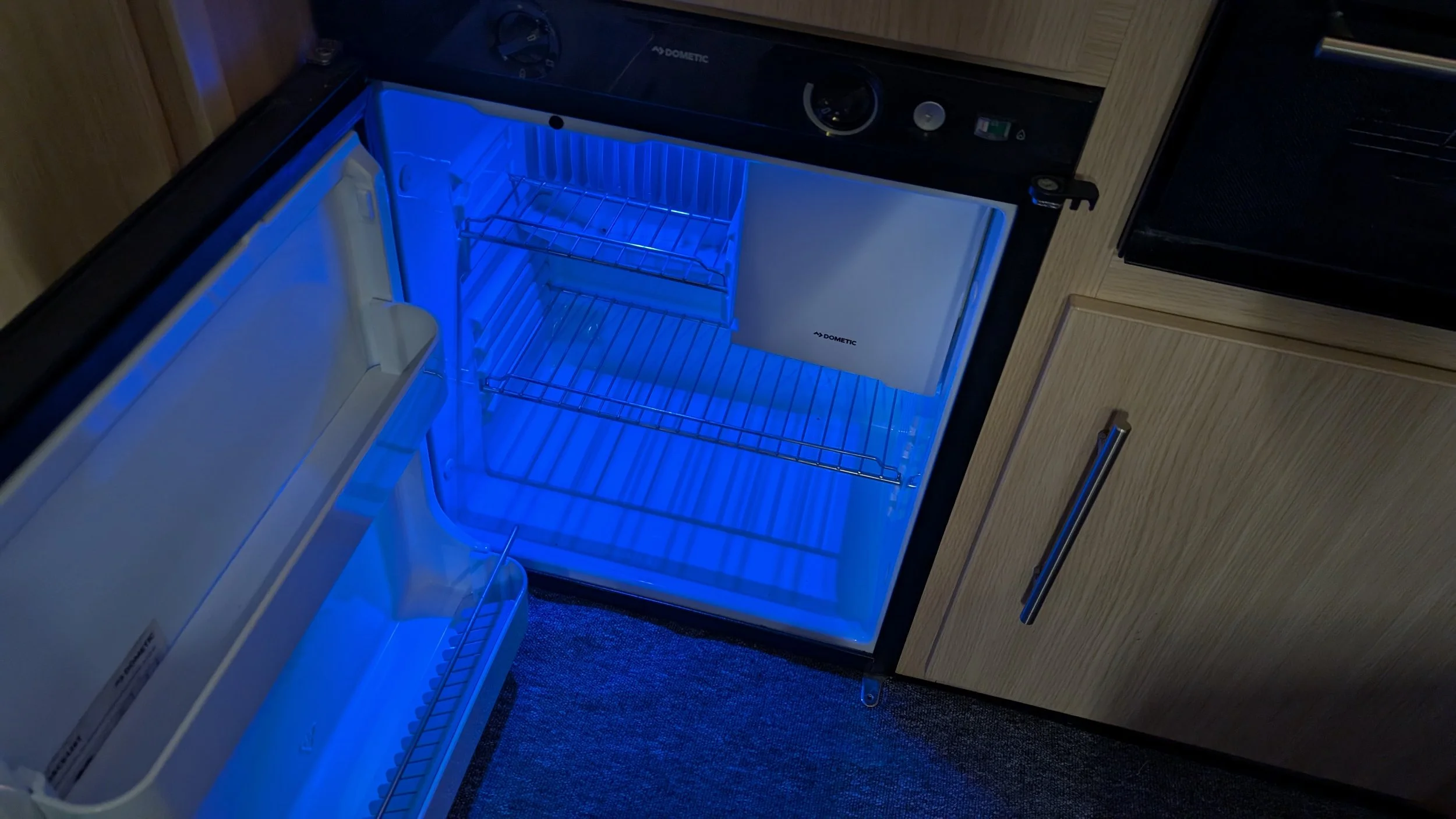An open refrigerator with blue LED lighting, located in a kitchen with wooden cabinets. The fridge is empty except for the shelves and baskets inside.