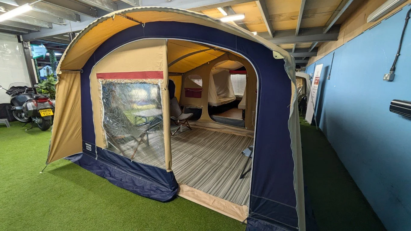 A camping tent displayed indoors, set up on artificial grass with a partially open flap revealing interior seating and flooring.