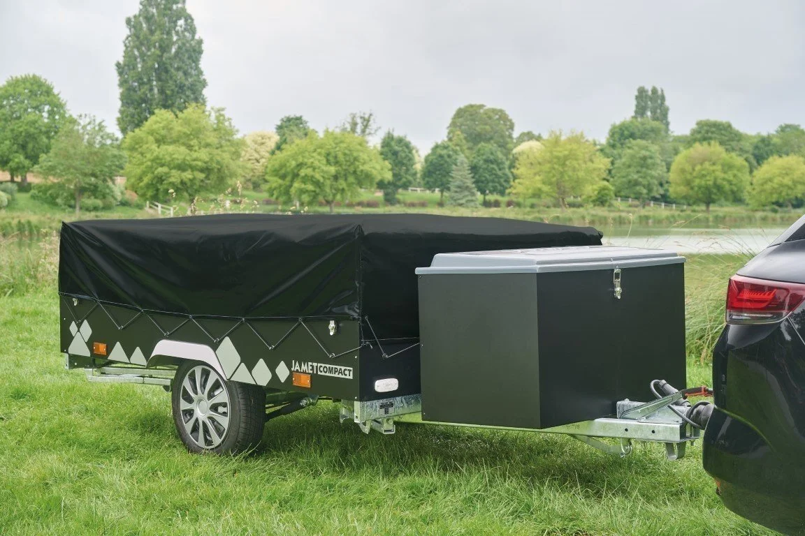 A black Jamet Compact trailer hitched to a black vehicle parked on grass near a river, with trees and cloudy sky in the background.