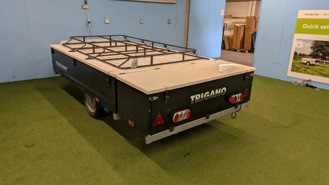 A black and white portable camping trailer with a mounted gas stove on top, sitting on a green artificial grass surface in an indoor setting.
