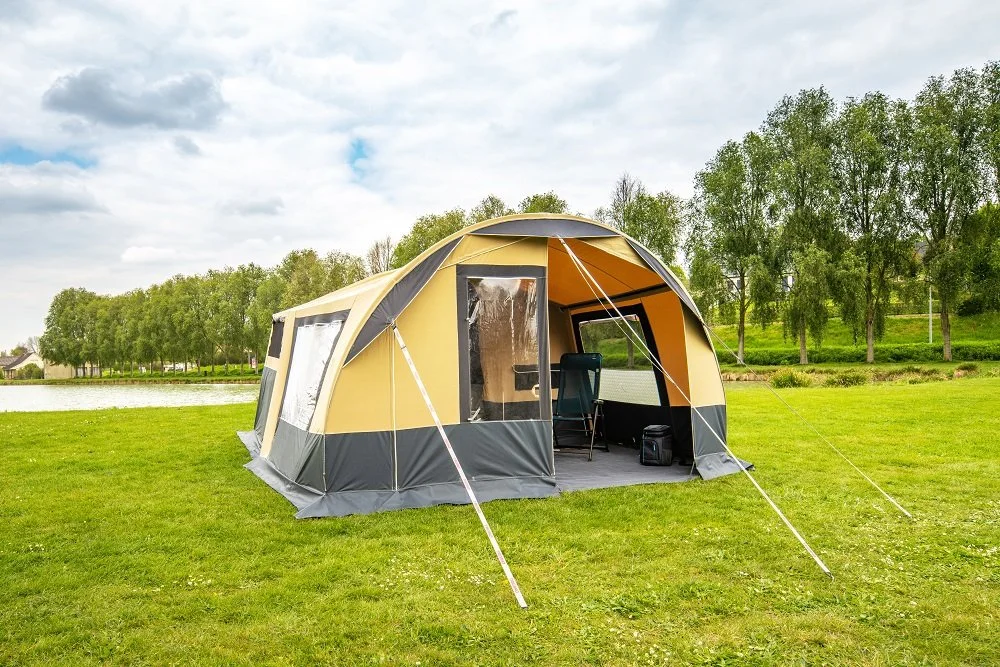 A large beige and gray Camplair tent set up on a grassy field near a lake, with trees in the background and cloudy skies overhead.