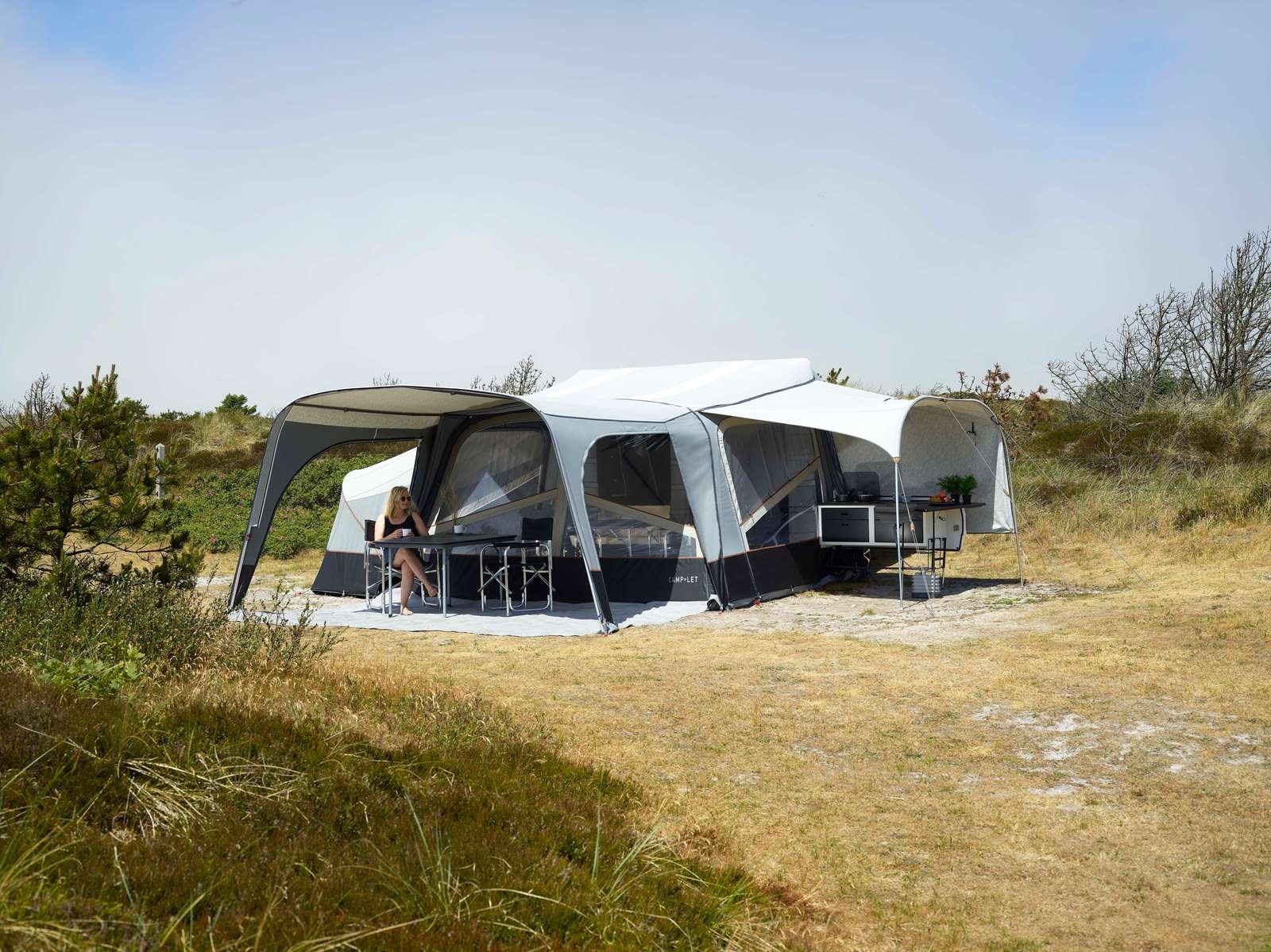 A woman sitting at a table outside a large, modern Camp-let tent in a grassy, open area with sparse trees, under a blue sky.