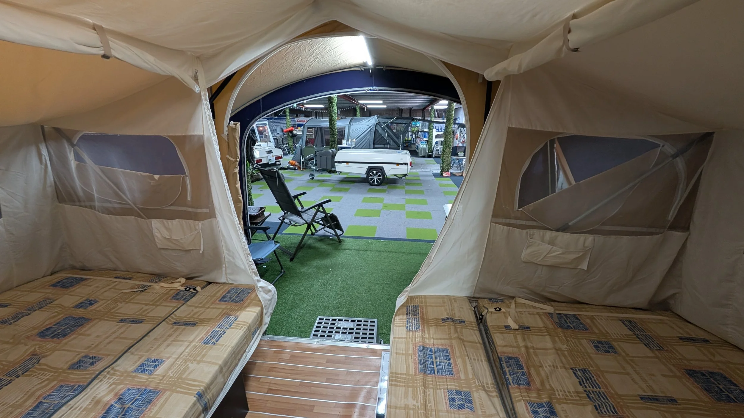 View from inside a camping tent looking out to an outdoor camping area with portable trailers, chairs, and artificial grass flooring.