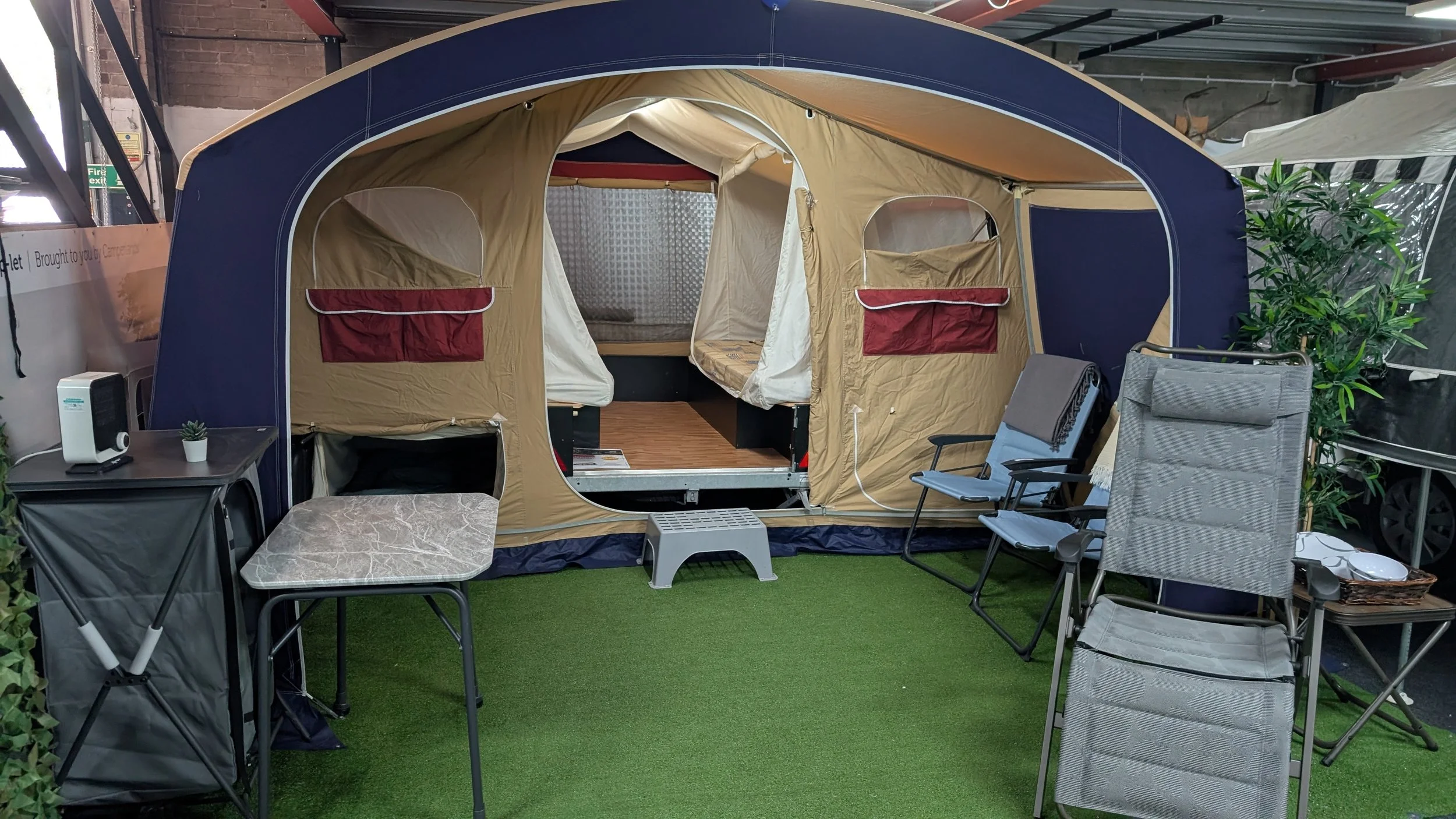 A camping tent set up indoors with various chairs and tables around it, and some potted plants. The tent has open windows and a door, with a platform inside and artificial grass on the floor.