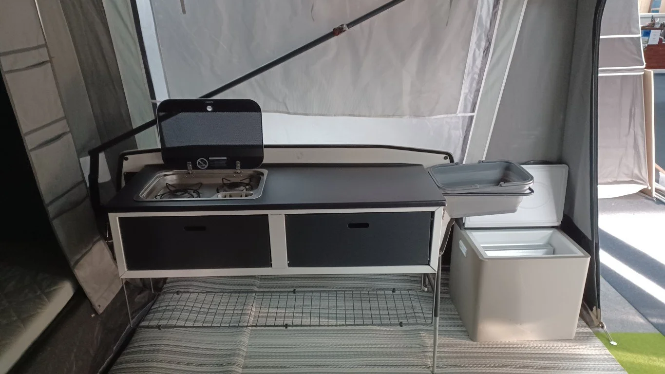 Portable outdoor kitchen setup with a small sink, two drawers, a countertop, and a mini refrigerator or cooler, all set inside a Camp-let Trailer Tent
