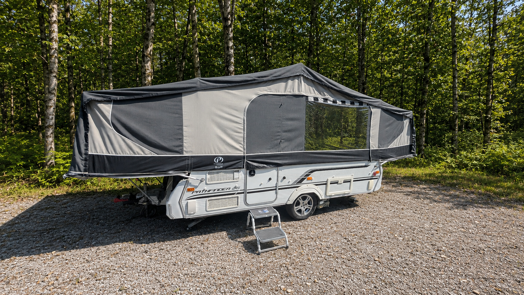 A white travel trailer with a pop-up tent attached, parked on a gravel surface in front of a dense forest with green trees.