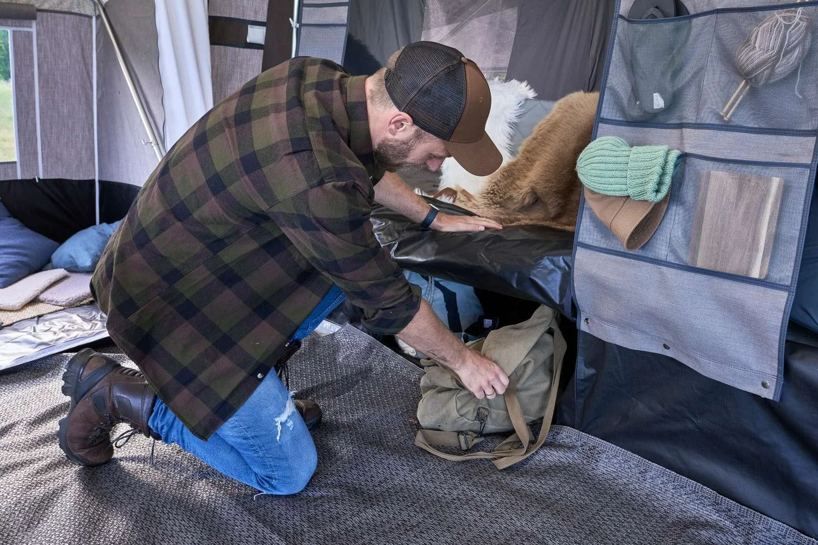 A man wearing a plaid shirt, jeans, and a cap kneels down and packs a backpack inside a tent. The tent has various camping items including a blanket, pillows, and a fur blanket. A hat and knitted glove are hanging on a mesh pocket.