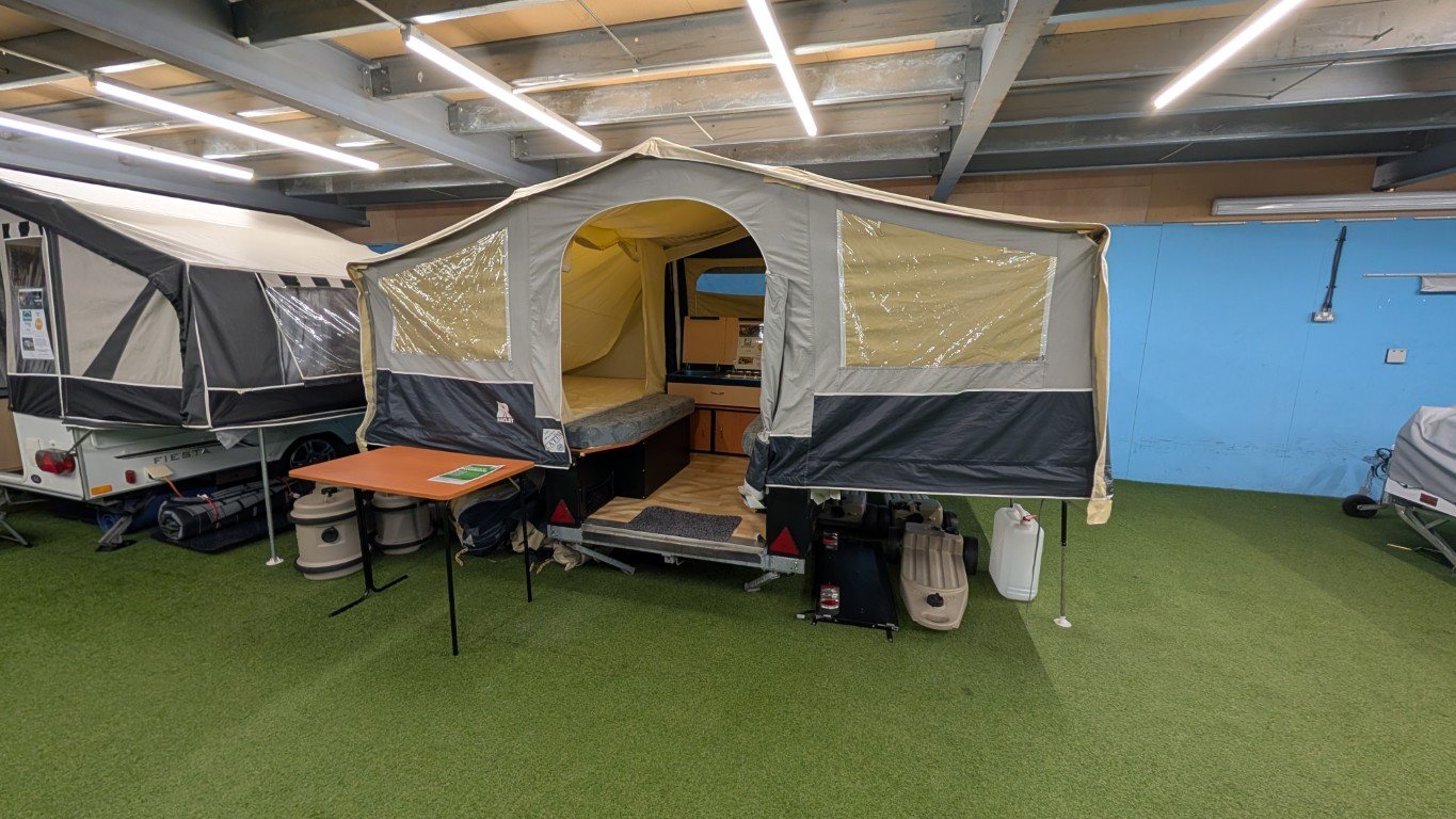 A camping trailer with a tent attachment inside an indoor showroom. The setup includes a small table, storage containers, and camping gear on artificial grass.