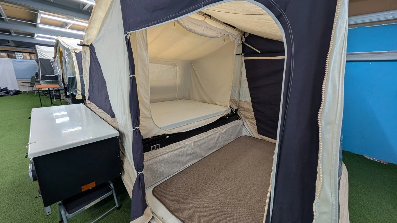 A pop-up camping tent set up indoors with a mattress inside, next to a white table or small fridge, on artificial green turf flooring.