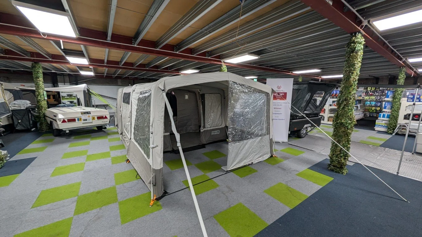 A large outdoor camping tent and a small camper trailer set up inside an indoor store. The tent has transparent plastic windows and is secured with white ropes. The store has a bright ceiling with multiple rectangular lights, carpeted flooring with g