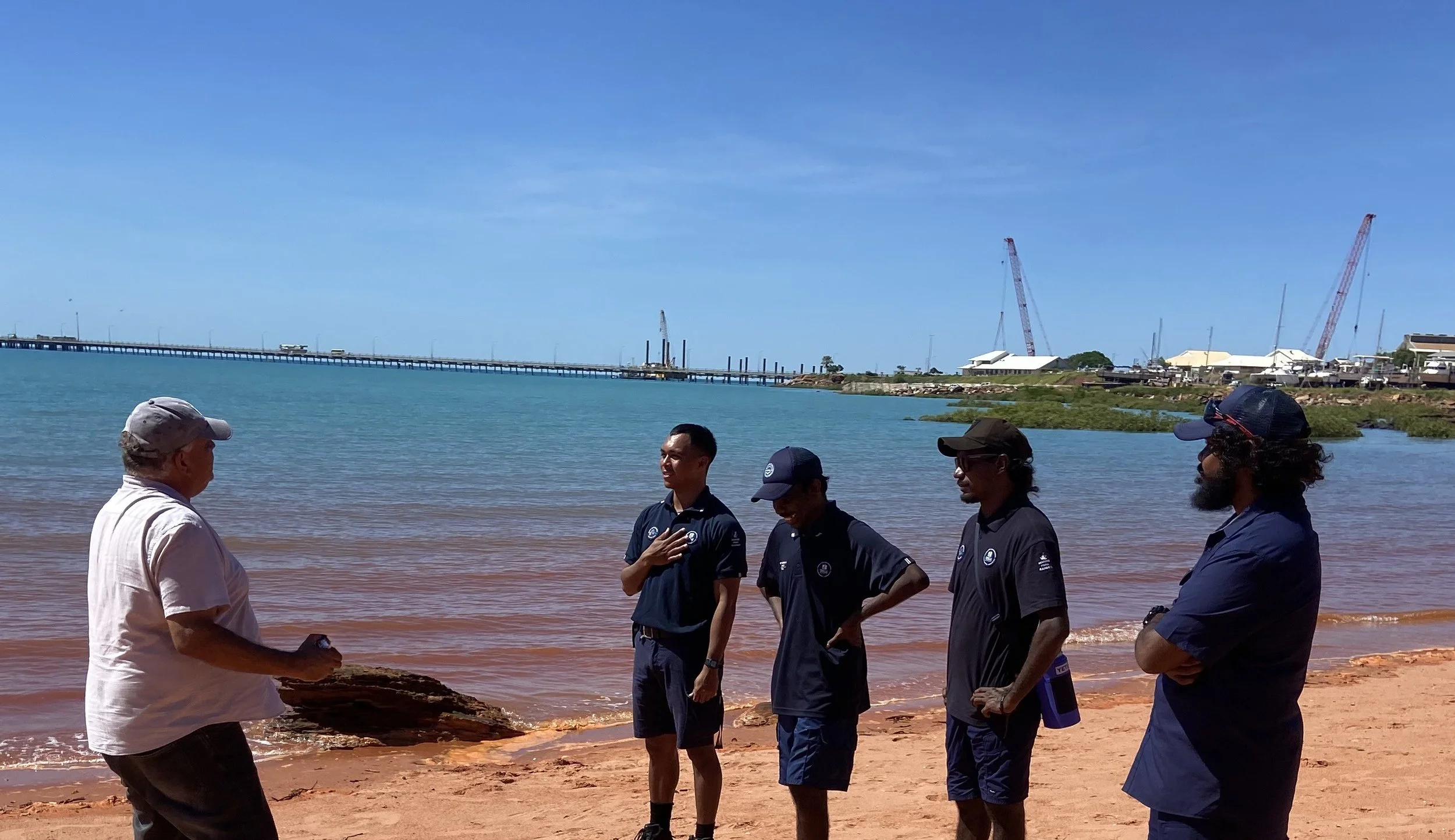 A group of five men standing on a sandy beach near the water, engaging in conversation, with a bridge, construction cranes, and buildings in the background under a clear blue sky.