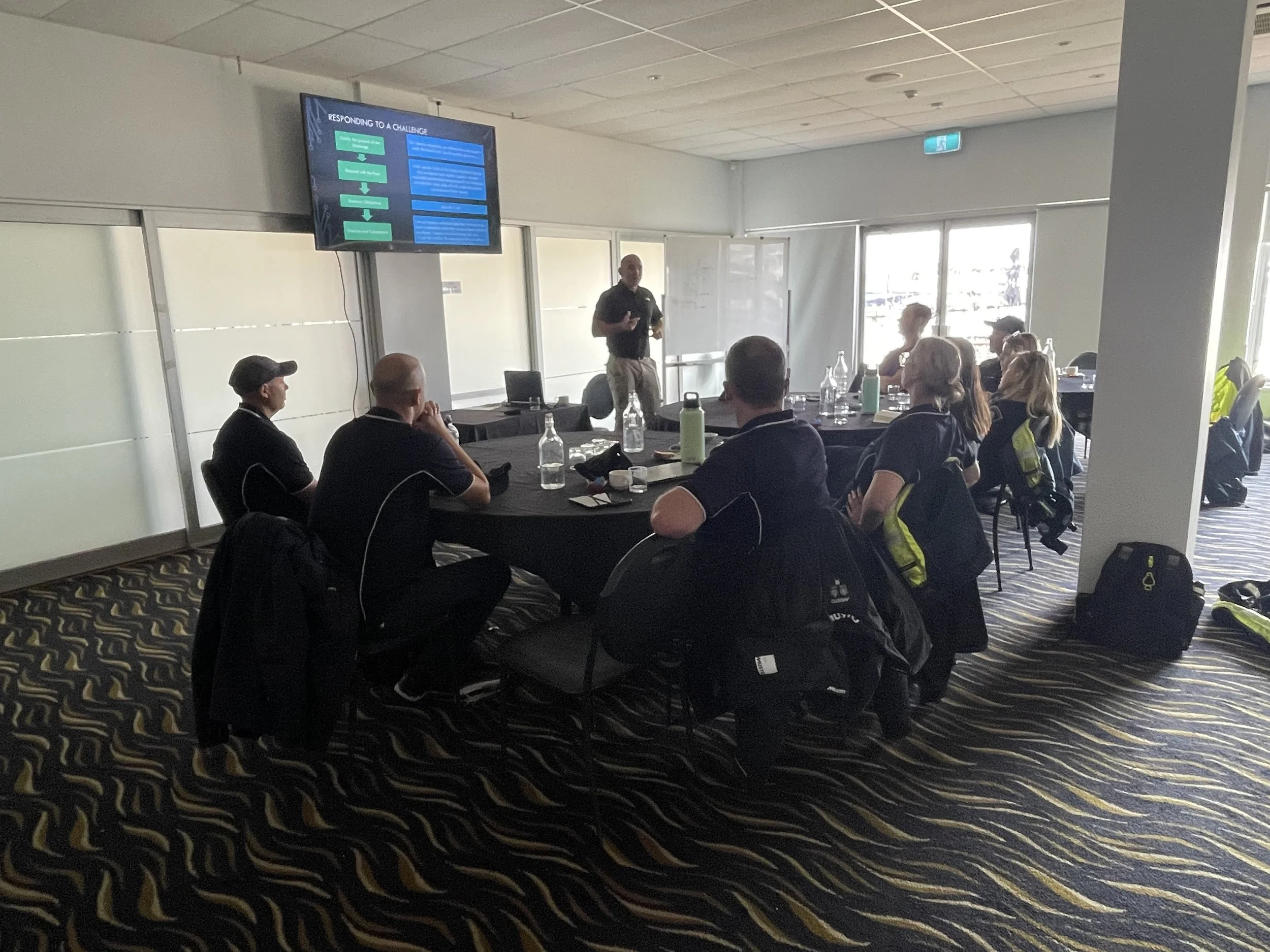 A group of people attending a presentation in a conference room, with a man standing and speaking in front of a screen displaying a slide titled 'Responding to a Challenge.'