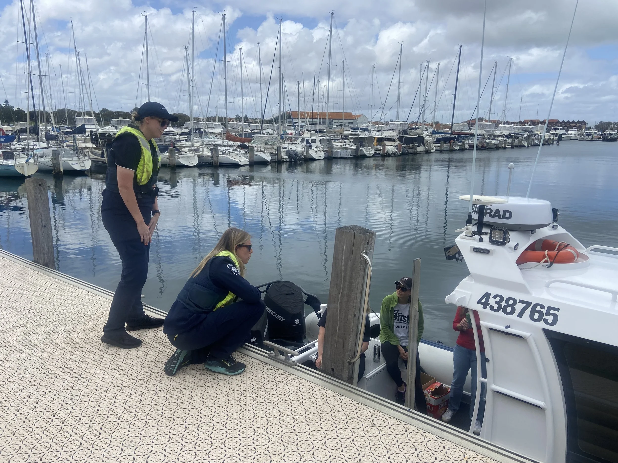 A group of people are working on a boat at a marina with sailboats docked and cloudy skies in the background.