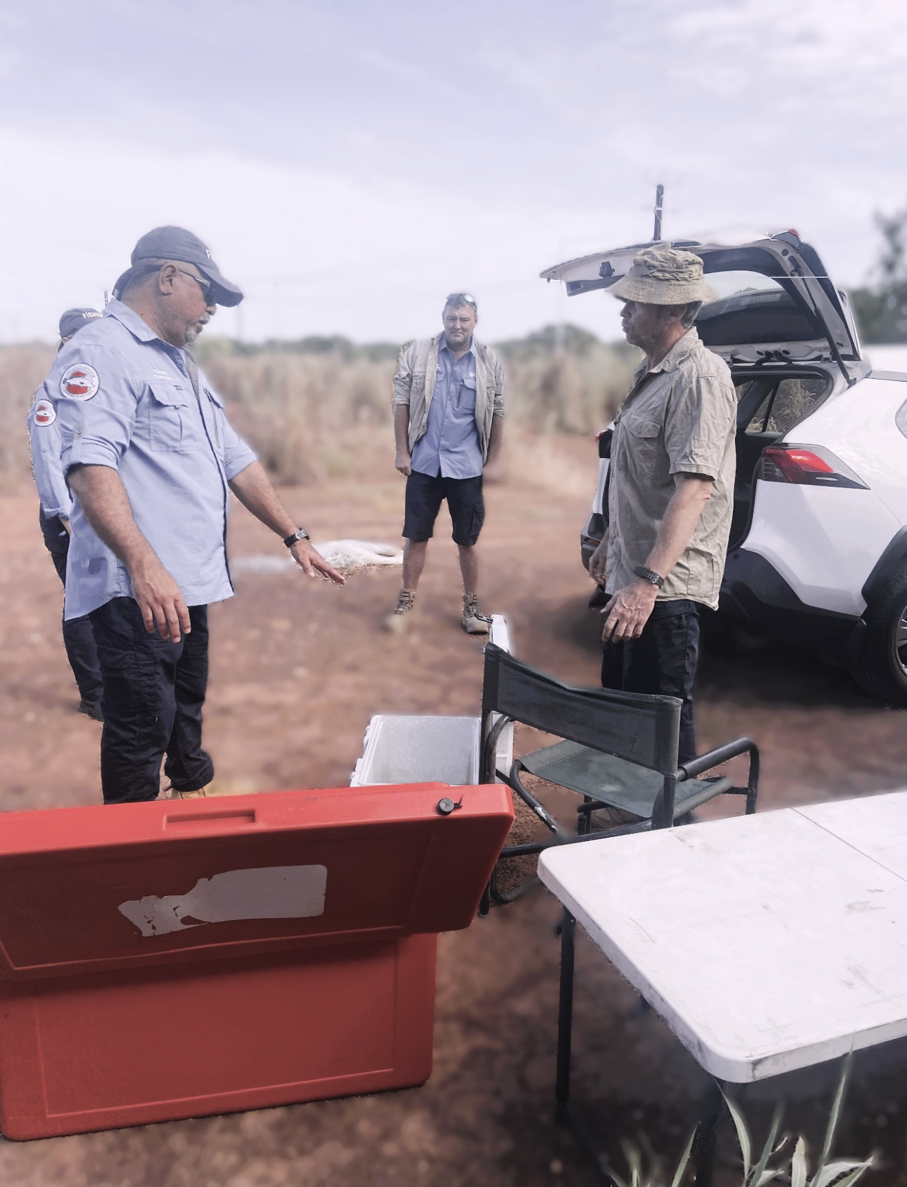 Four men outdoors around a table with equipment, one man holding a fish, a car with open trunk, and a red container in the foreground.