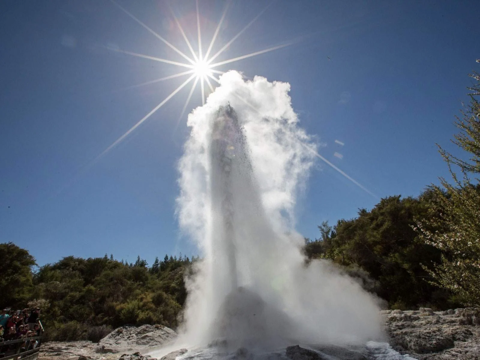 A geyser eruption with water shooting high into the air, sunlight radiating from above, and a clear blue sky in the background. There are trees on the horizon and people observing from the side.