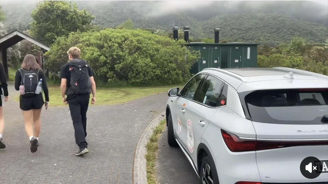 Three people with backpacks walking on a paved path near a white car parked along the side. There is greenery, bushes, and trees in the background with a mountain partially obscured by fog.