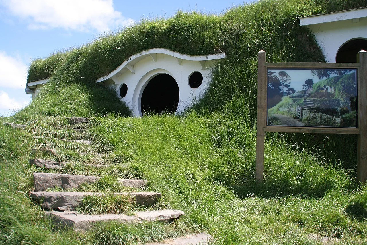 A hobbit house with a grass-covered roof, circular windows, and a stone staircase leading to the door, surrounded by green grass and trees.