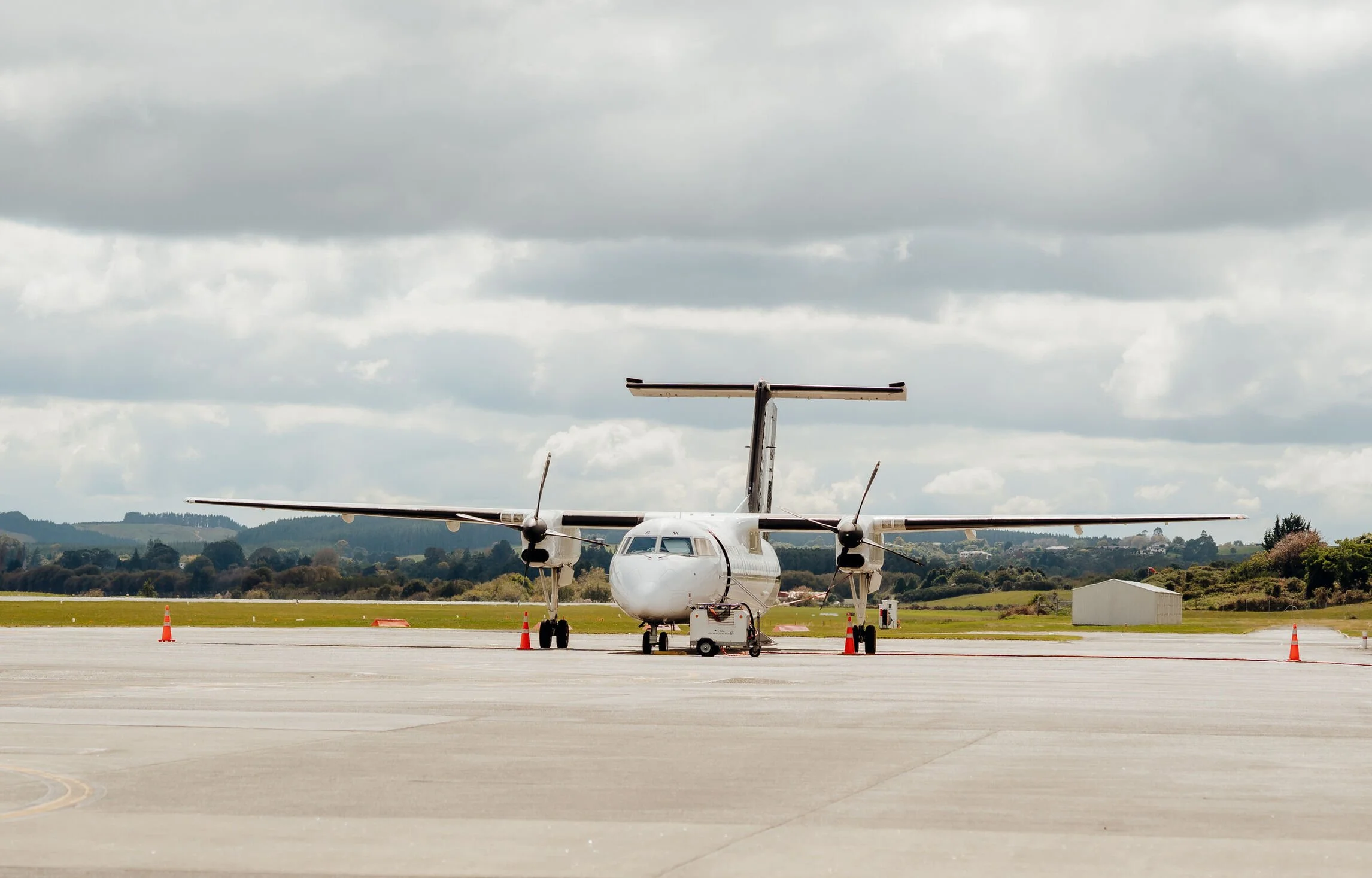 Small white propeller airplane parked on runway with orange cones around it, cloudy skies above, and trees in the background.