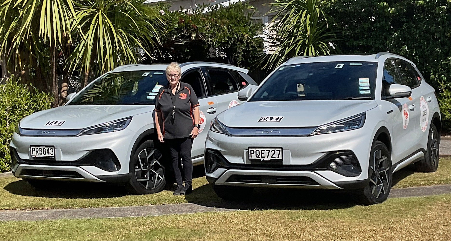 A woman standing between two white taxis on a grassy area, with trees and plants in the background.