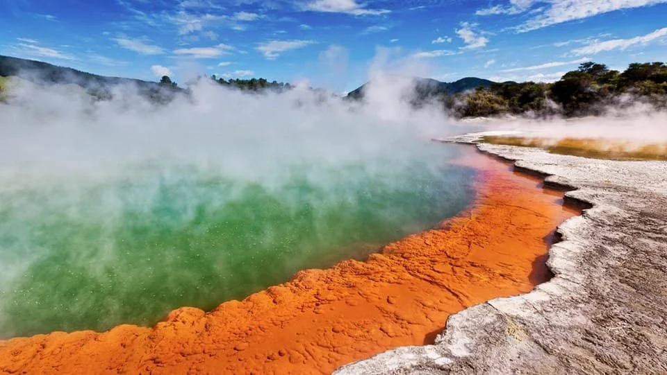 Colorful geothermal hot spring with green water, bright orange mineral deposits, steaming surface, in a scenic outdoor landscape with hills and cloudy sky.