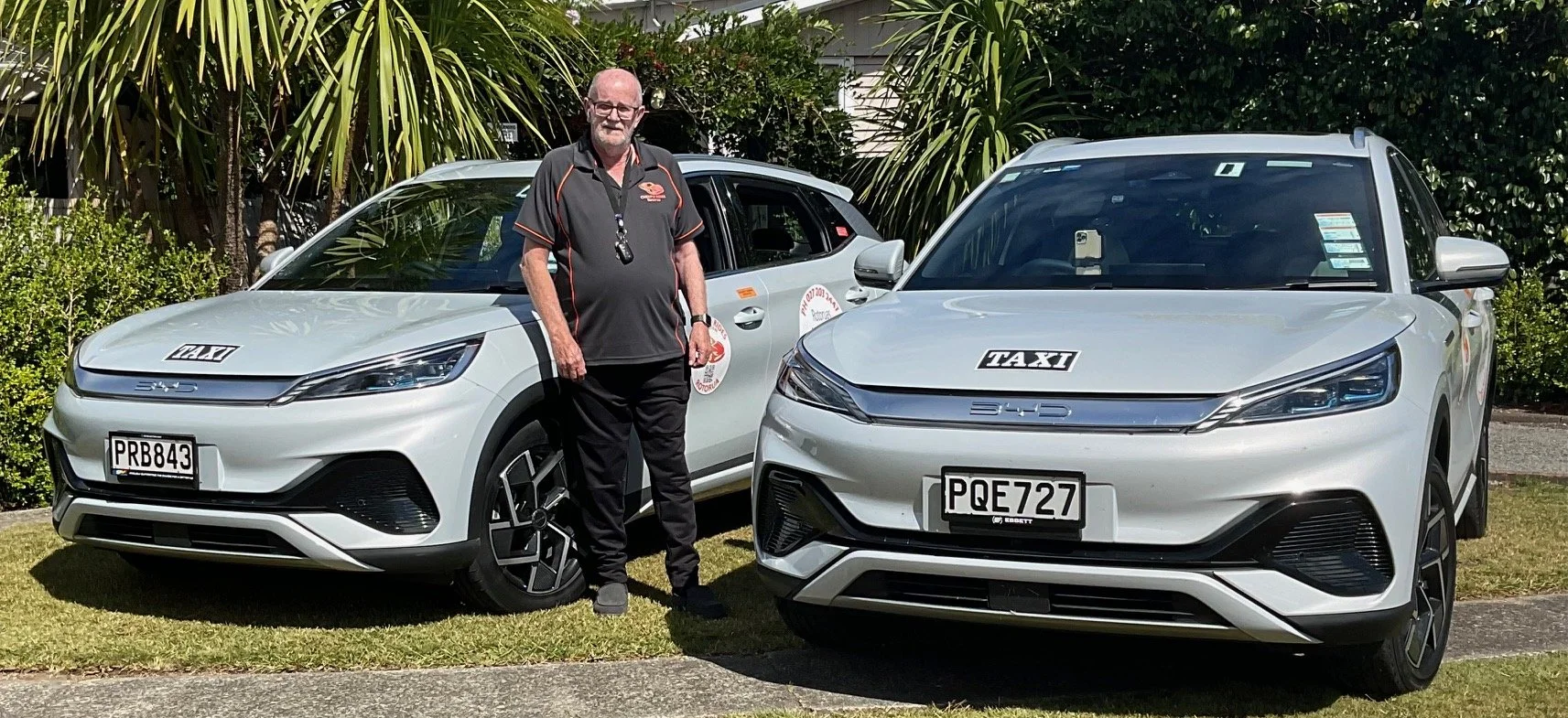 A man in a black shirt standing between two light gray electric vehicles with taxi signs, parked on a grassy area with trees and bushes in the background.