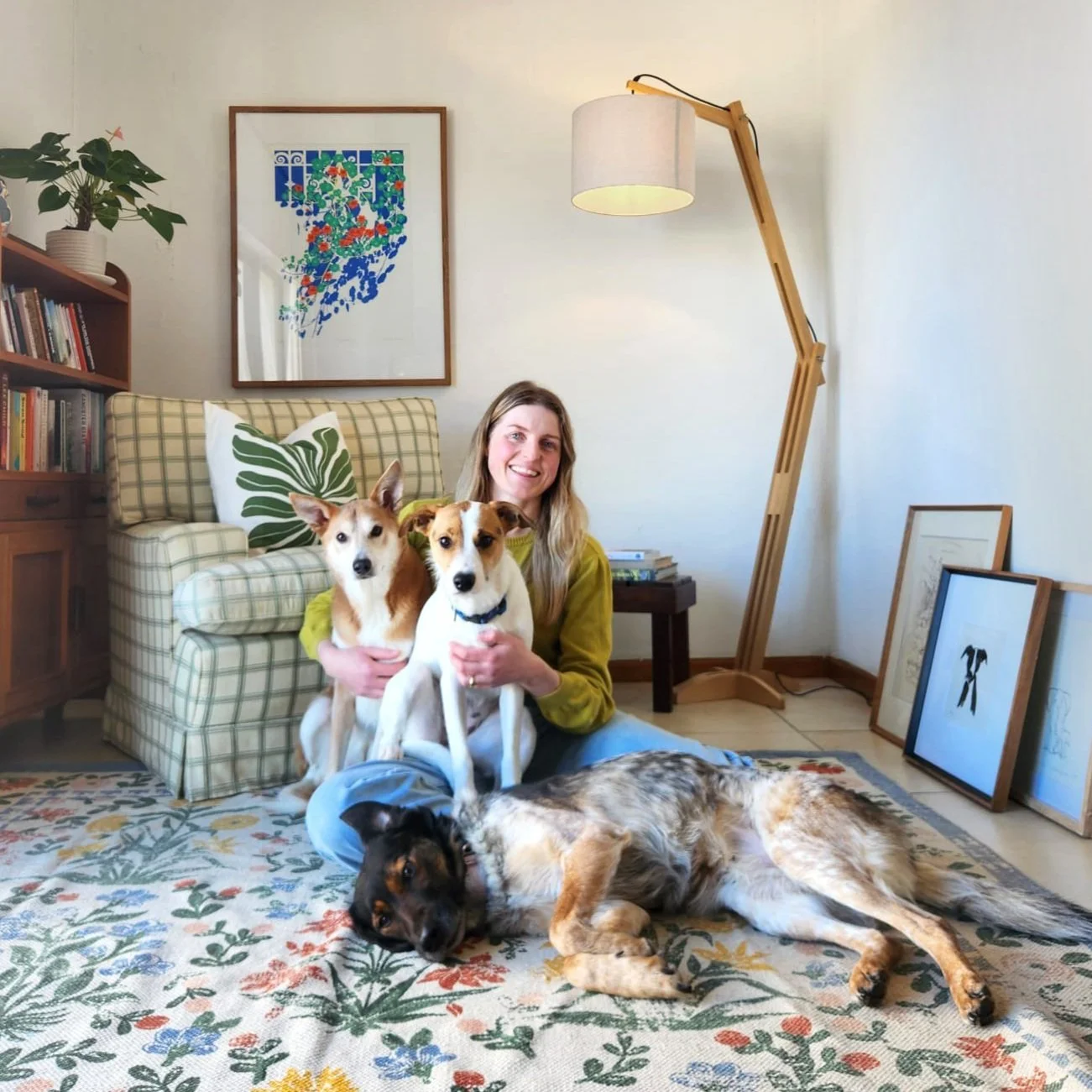A woman sitting on the floor with three dogs in a cozy living room. One of the dogs is sitting on her lap, another in front of her, and a third lying on the carpet in front. The background features a bookshelf, framed artwork, and a floor lamp.