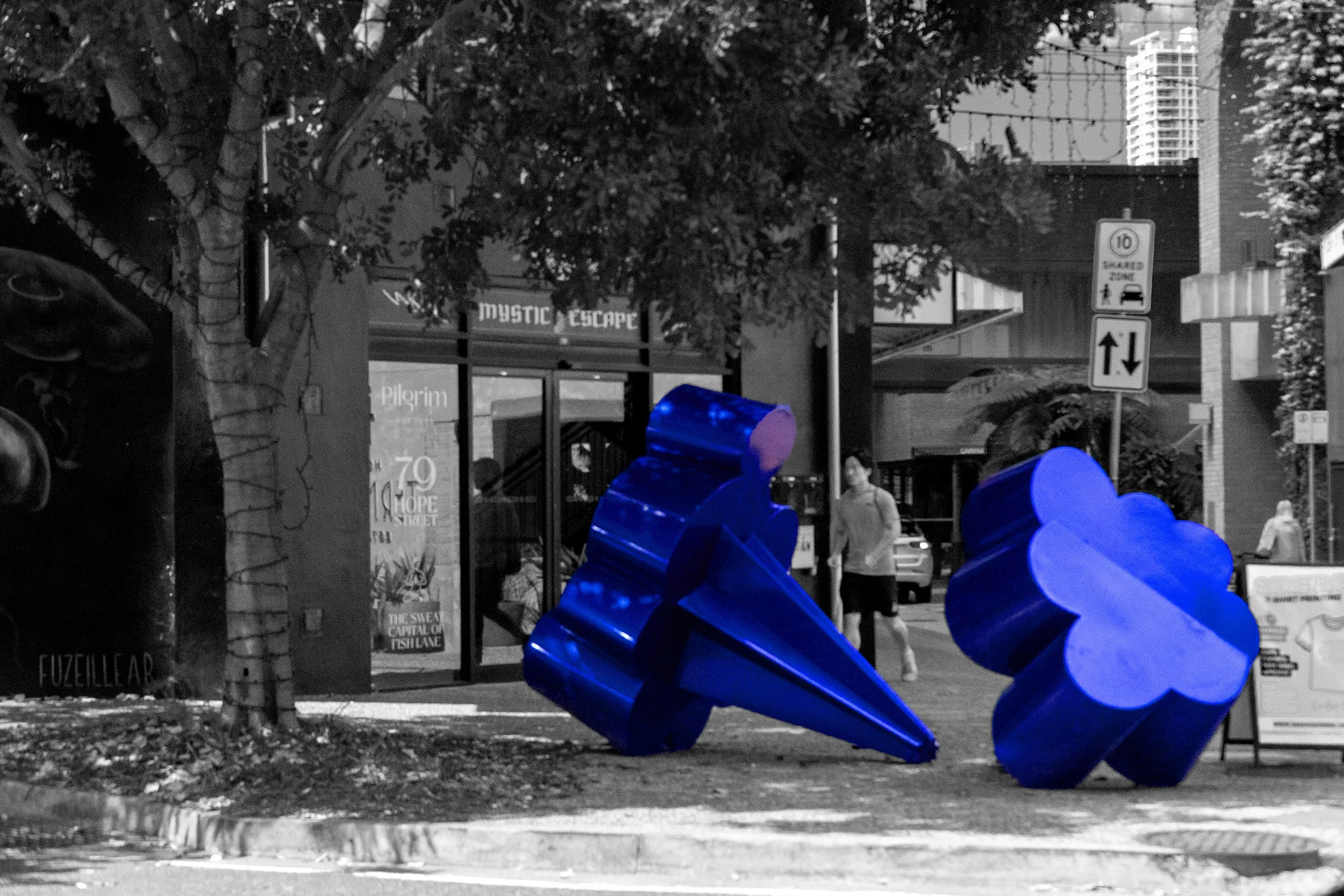 This photograph highlights a vibrant blue sculpture against a monochromatic urban background. The selective colour technique draws attention to the artwork's unusual shapes, creating a striking contrast between public art and its surroundings.