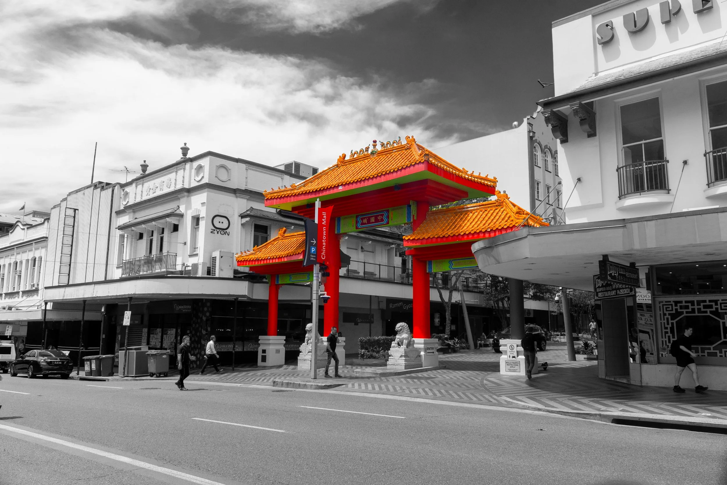 This photograph captures a traditional Chinese gateway surrounded by monochromatic urban buildings. The vibrant orange roof and red pillars become the focal point through selective colour, highlighting the contrast between cultural heritage and the m