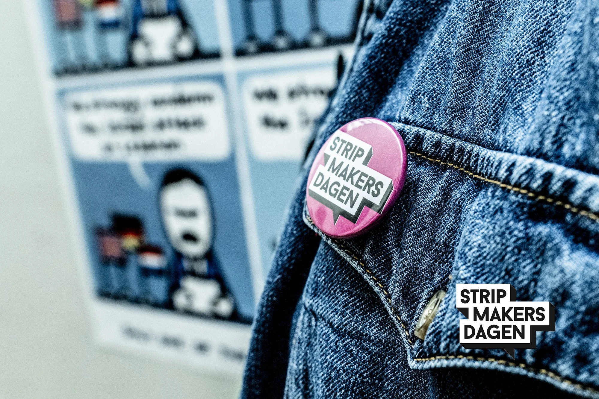 Een close-up van een denim jas met een roze knoop met tekst 'Strip Makers Dagen'.