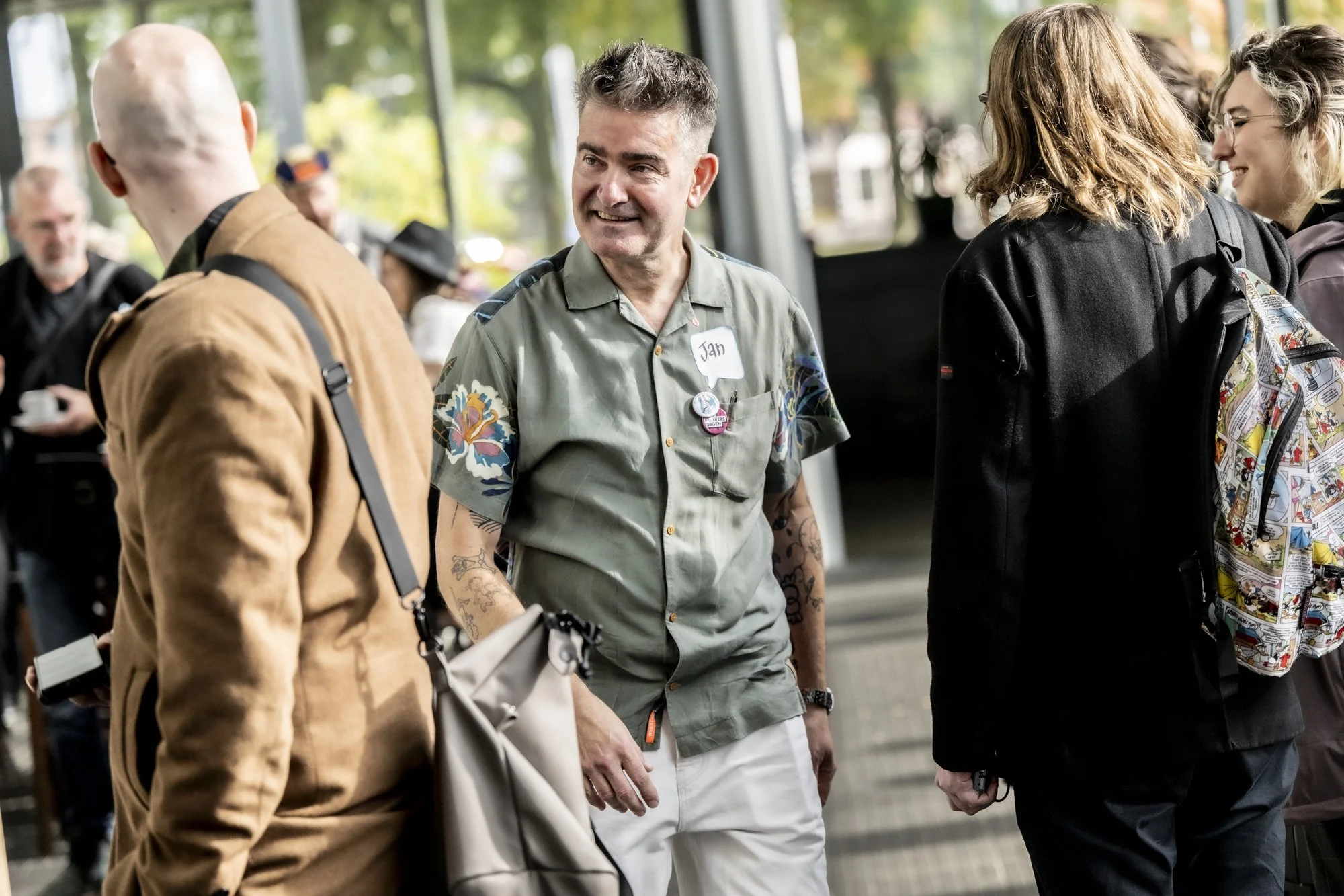 Mann met donkere haar en een tatoeage op zijn armen, lachend en in gesprek met andere mensen in een openbare ruimte, omgeven door meerdere mensen die op een informele manier met elkaar praten.