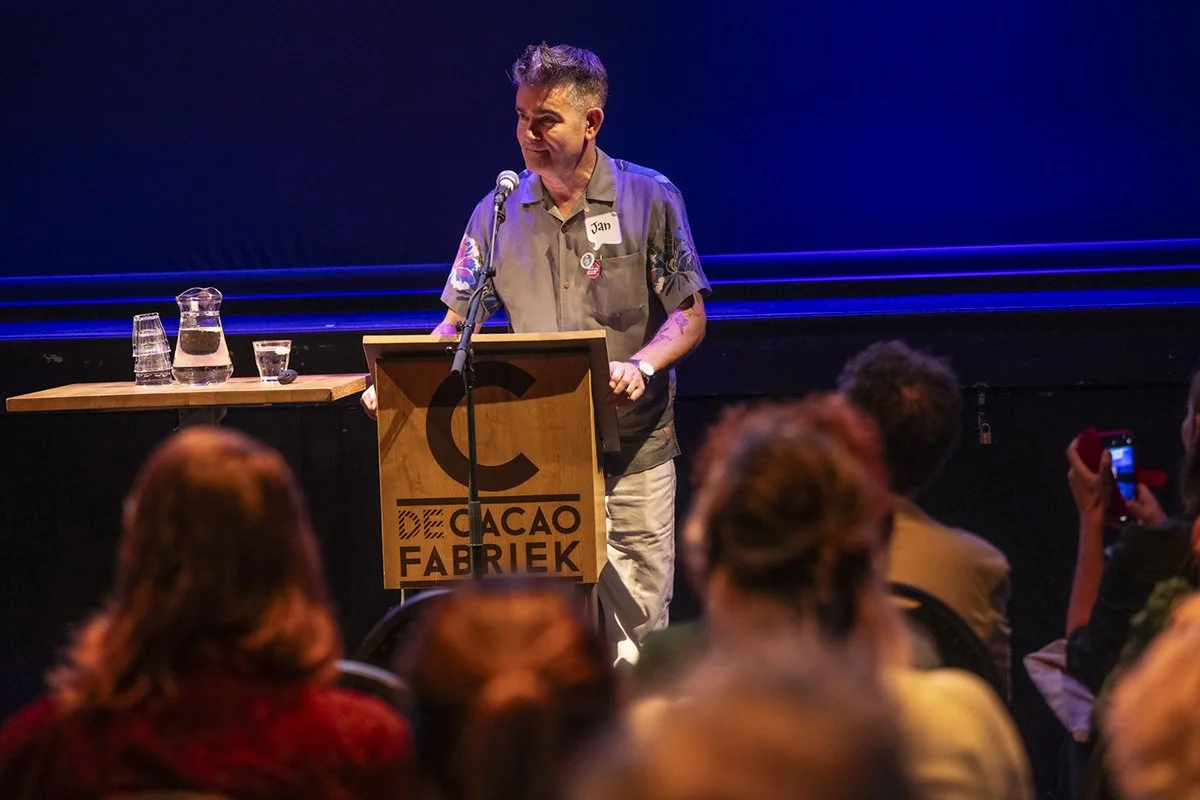 Jan Vriends geeft een presentatie achter een houten spreekgestoelte met het logo 'De cacao fabriek' en een publiek kijkt toe, het decor is donker met blauwe verlichting.